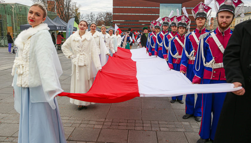 Parada Niepodległości we Wrocławiu. Zdjęcie archiwalne. Widać na nim kobiety (po lewej stronie) i mężczyzn (po prawej) ubranych w stroje historyczne, którzy trzymają polską flagę