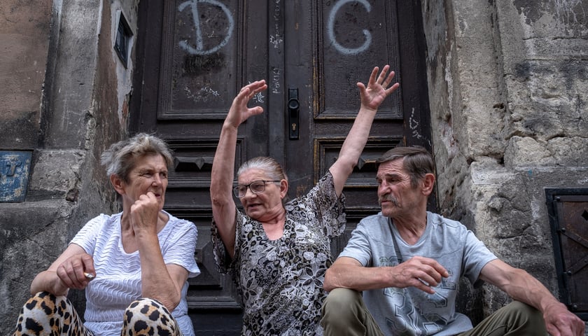Diw kobiety i mężczyzna siedzą przed popisanymi drzwiami kamienicy w Legnicy, jedna z kobiet unosi ręce w ekspresyjnym geście
