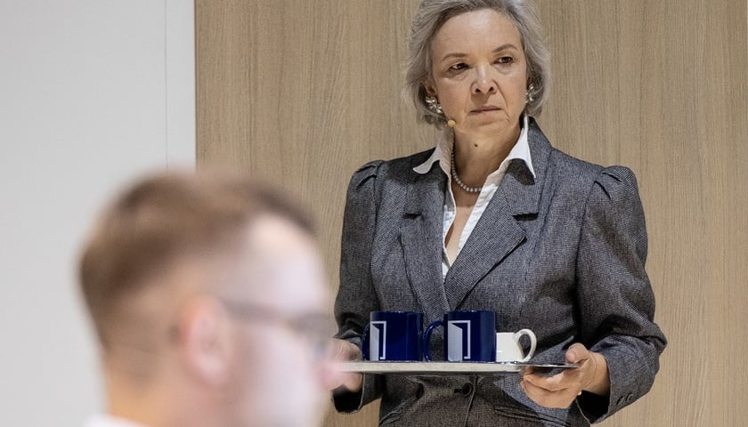 Aktorka Zina Kerste, ubrana w grafitową marynarkę, niesie tackę z kubkami (zdjęcie z próby)