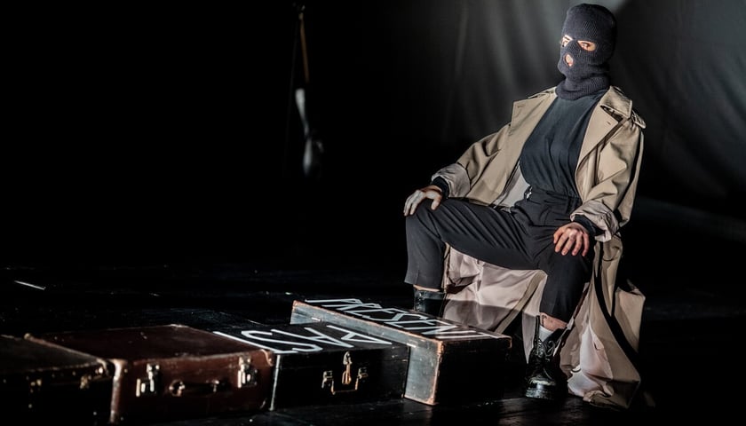 Spektakl teatralny "Niesamowita Słowiańszczyzna". Na zdjęciu kobieta w czarnej kominiarce siedząca na krześle. Na lewo od niej rząd podróżnych walizek.
