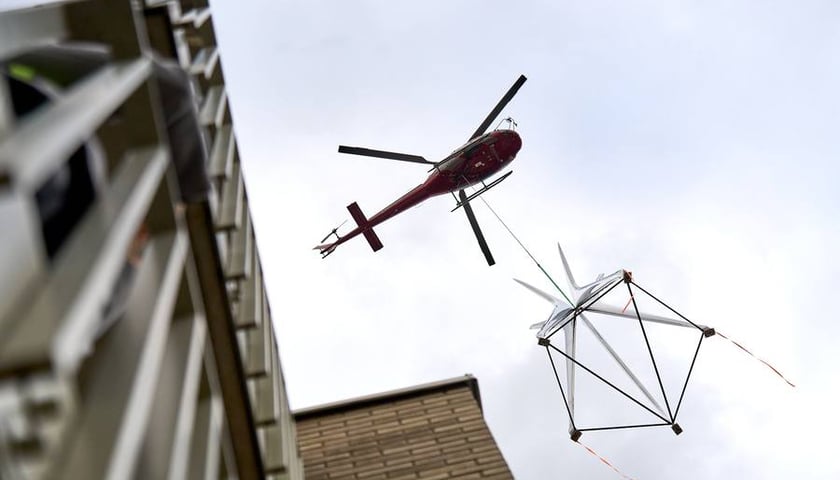 Rzeźba podwieszona na linie do helikoptera podczas lotu, widać też kawałek budynku