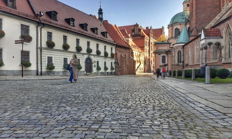 Na pierwszym planie jest kostka brukowa Placu Katedralnego (spaceruje po nim kobieta), w XIV wieku był tu cmentarz parafii diecezjalnej na Wyspie Tumskiej