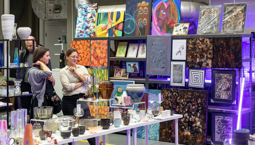 Dwie dziewczyny przy stole, na którym stoją m.in. wyroby szklane i ceramiczne, widać także obrazy (budynek ASP Wrocław przy Traugutta)