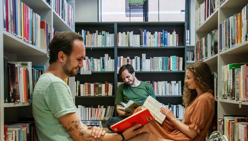 3 osoby (2 mężczyzn i 1 kobieta) siedzące na podłodze i czytające książki. W tle biblioteczne półki
