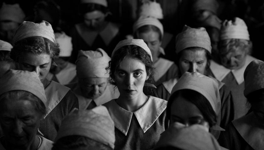 Kadr z filmu "Dziewczyna z igłą". Czarno-białe zdjęcie przedstawiające młodą kobietę o intensywnym spojrzeniu patrzącą w obiektyw. Wokół niej wiele innych kobiet z opuszczonymi głowami.