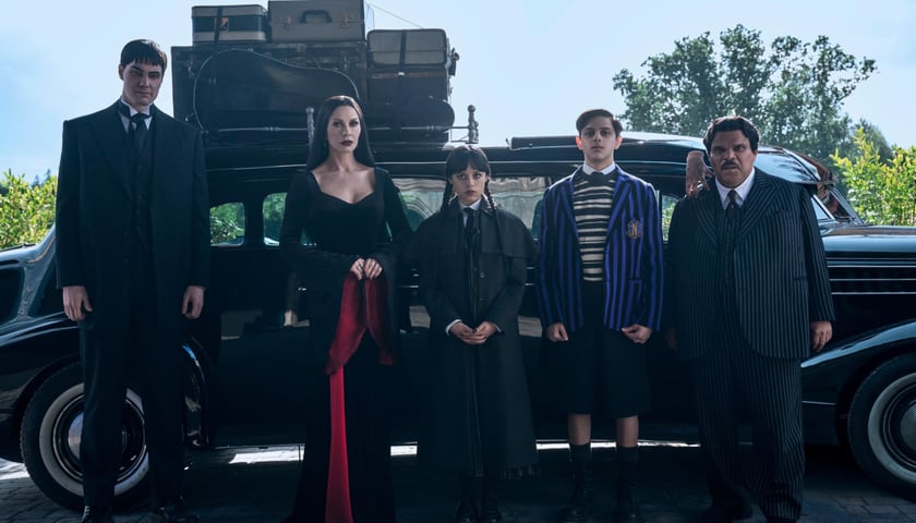 Kadr z serialu „Wednesday” Netflixa: 5 osób stoi przed czarnym samochodem, w środku Jenna Ortega (tytułowa bohaterka), obok niej z prawej Isaac Ordonez (Ajax), który będzie gościem spotkania we Wrocławiu