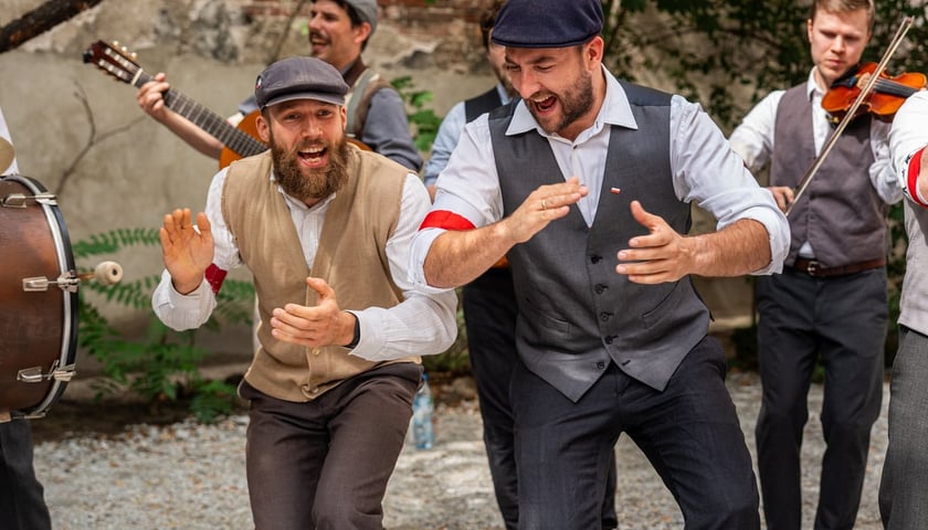 Koncert Ferajny Hoovera. Na pierwszym planie dwóch mężczyzn w kaszkietach z biało-czerwonymi opaskami na przedramieniu 