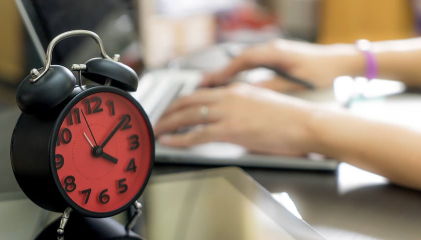 Na zdjęciu budzik i osoba pisząca na laptopie w biurze