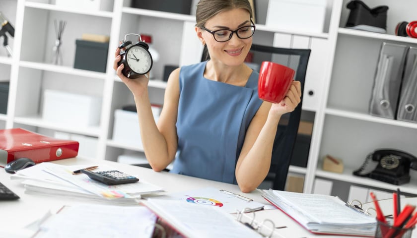 Na zdjęciu kobieta w biurze z kubkiem kawy i zegarkiem w rękach / zdjęcie ilustracyjne