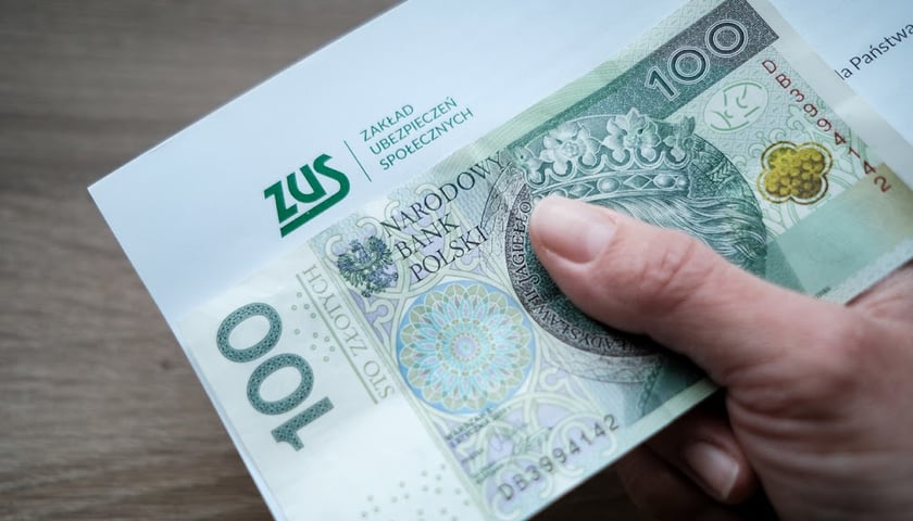 Na zdjęciu widać dłoń, banknoty i kopertę z nadrukiem ZUS 