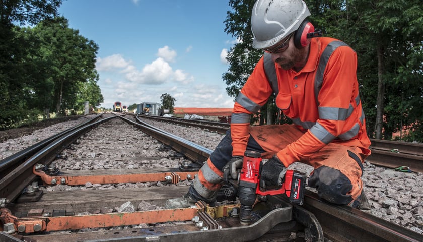 Robotnik w kasku i pomarańczowym kombinezonie pracujący na torach kolejowych, w rękach ma narzędzie Milwaukee