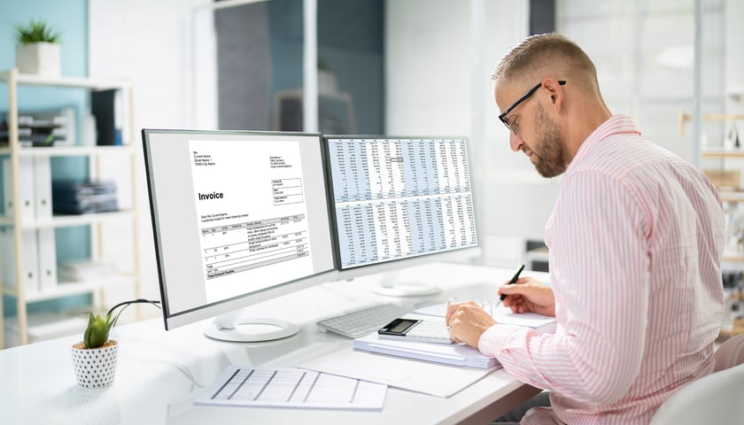 Mężczyzna przegląda dokumenty i liczy na kalkulatorze na biurku, w tle monitory komputerów