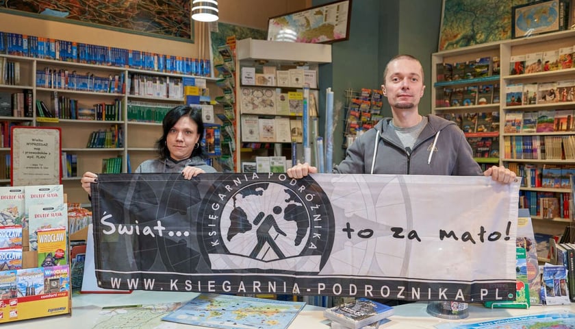 Pracownicy Księgarni Podróżnika trzymają baner z napisem "Świat to za mało.. Księgarnia Podróżnika", w tle regały z książkami i mapami