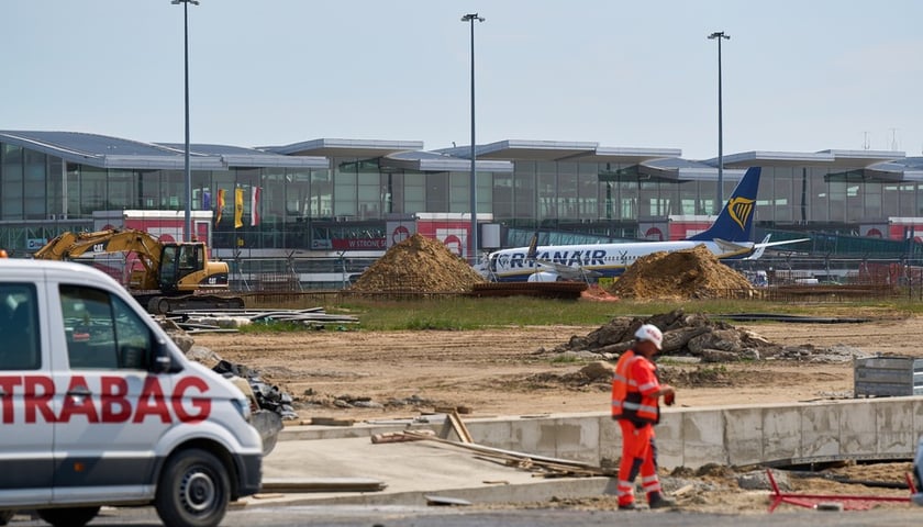 Na pierwszym planie jest plac budowy z robotnikiem, w głębi widać samolot i terminal wrocławskiego lotniska