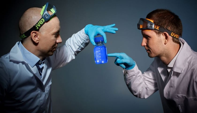 Dwóch naukowców (Artur Podhorodecki i Mateusz Bański, współzałożyciele QNA Technology S.A.), trzymających buteleczkę z niebieskim płynem (roztwór z kropkami kwantowymi) 