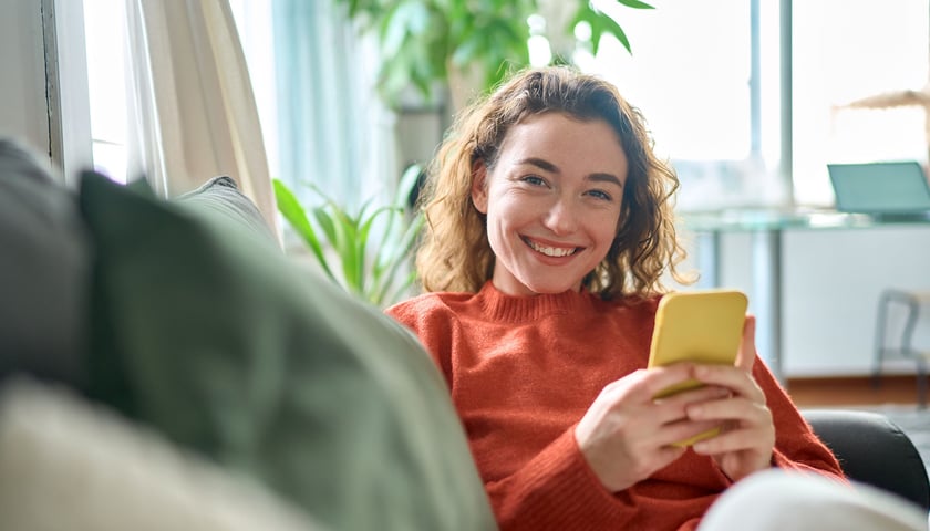Uśmiechnięta kobieta ze smartfonem w ręku, w tle widać kanapę, zdjęcie ilustracyjne