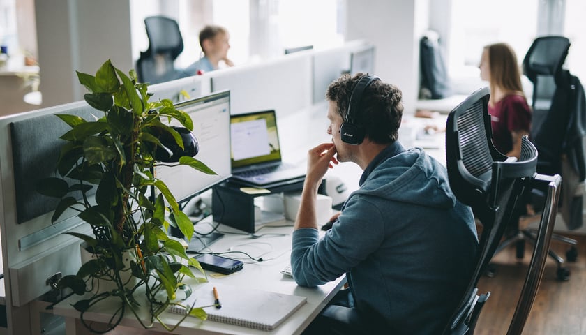 Mężczyzna w słuchawkach patrzy na monitory komputerów, w tle kobieta i monitory. Pracownicy spółki Monterail