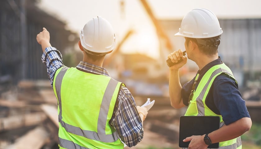 Dwóch mężczyzn w kaskach i kamizelkach ochronnych na budowie, w tle dźwig, zdjęcie ilustracyjne