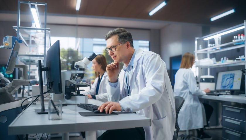 Mężczyzna w okularach, siedzi i patrzy na ekran monitora, w tle dwie kobiety w laboratorium, zdjęcie ilustracyjne 