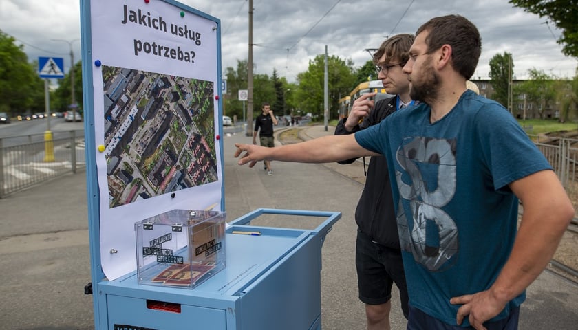 Powiększ obraz: Konsultacje w sprawie terenu TBS-u przy ul. Głubczyckiej we Wrocławiu. Na zdjęciu widać dwóch mężczyzn analizujących plan inwestycji zawieszony na wózku konsultacyjnym.