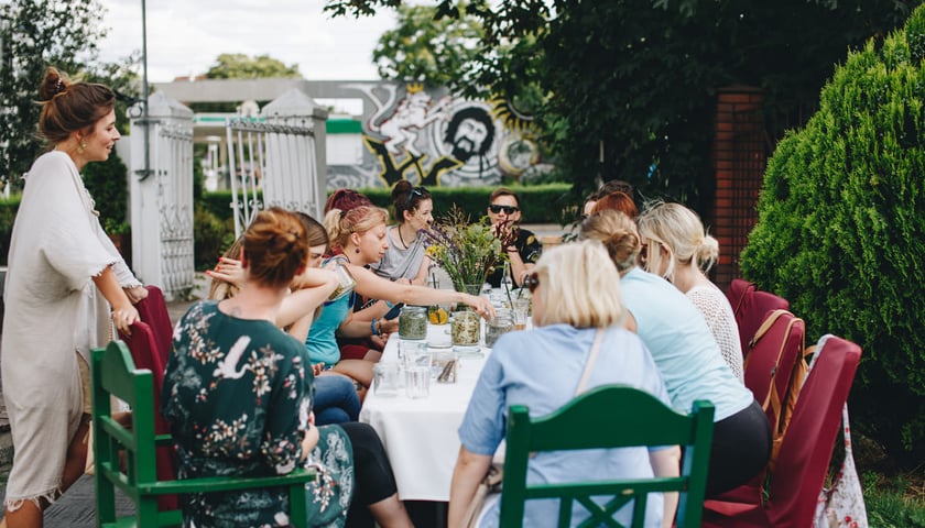 Mikrogranty, projekt "Wrocławskie Święto Kwiatów". Na zdjęciu podłużny stół ustawiony w plenerze, przy stole siedzą ludzie. Na stole kwiaty polne, szklanki z wodą, słoje z różną zawartością.