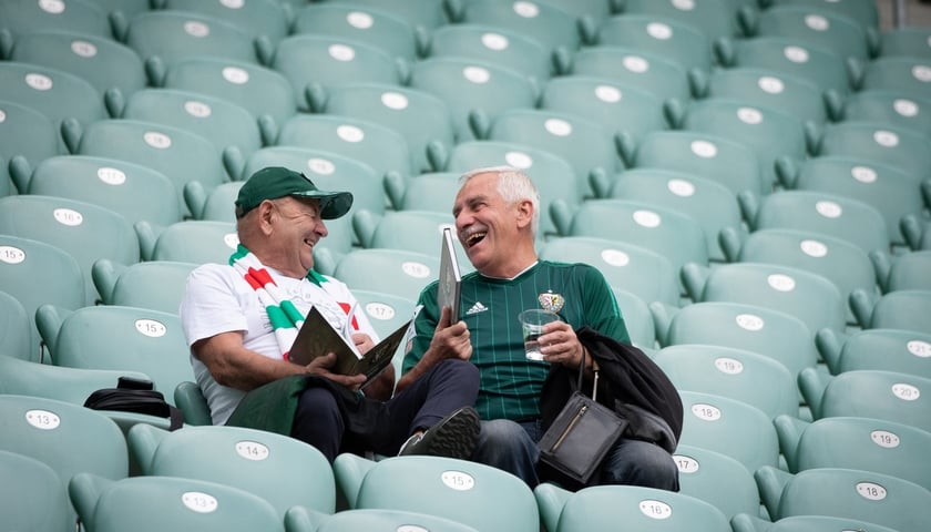 Powiększ obraz: na zdjęciu dwóch kibiców-seniorów na zielonych krzesełkach na trybunie stadionu