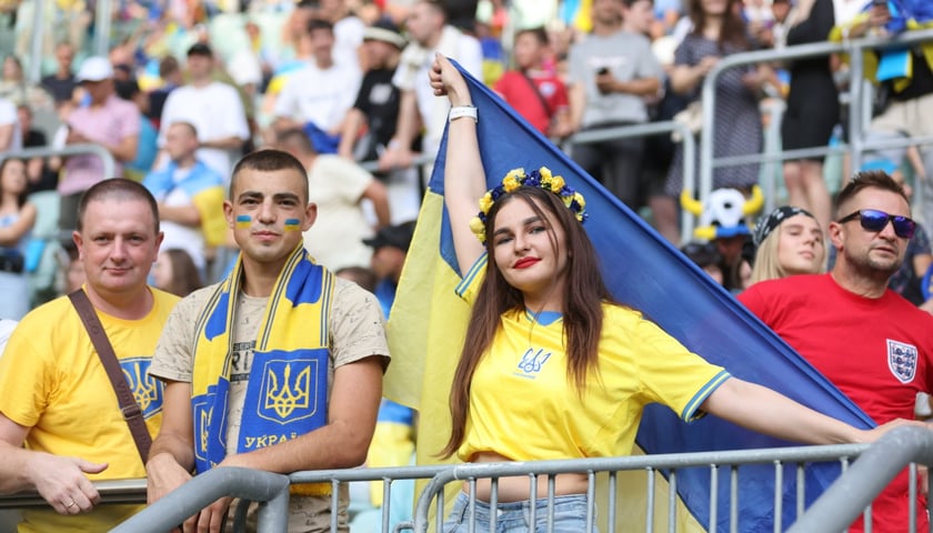 Powiększ obraz: Dwóch kibiców w narodowych barwach Ukrainy i kibicka z żółto-niebieską flagą i kwiatami we włosach