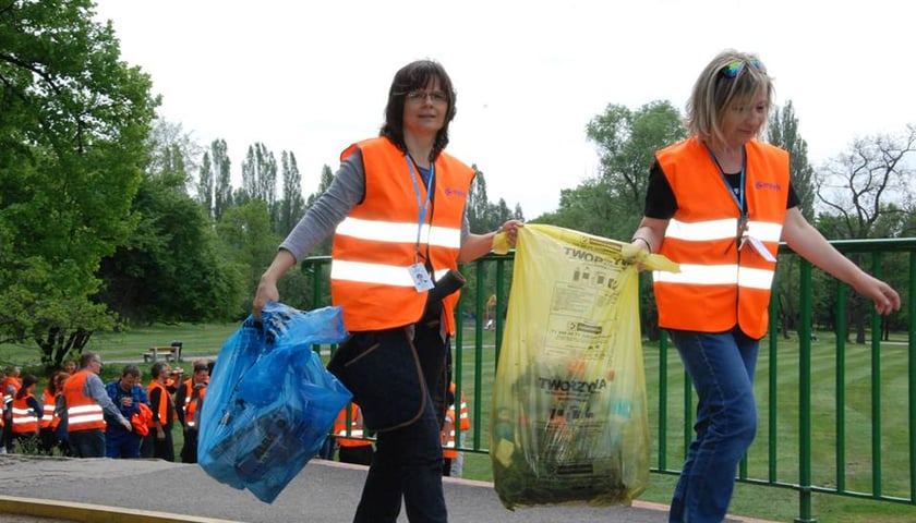 Powiększ obraz: Dwie kobiety niosą worki z odpadami. Noszą pomarańczowe kamizelki odblaskowe.