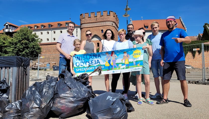 Na zdjęciu widać grupę ludzi, uczestników akcji, która sprzątała przez ostatnie trzy lata śmieci porozrzucane po Polsce. Pierwszy od prawej - Dominik Dobrowolski. Koło ludzi, przy ich nogach, leżą worki pełne śmieci, które zebrali. Ludzie widoczni na zdjęciu trzymają przed sobą baner z napisem Wiślana Odyseja, sprzątamy rzeki