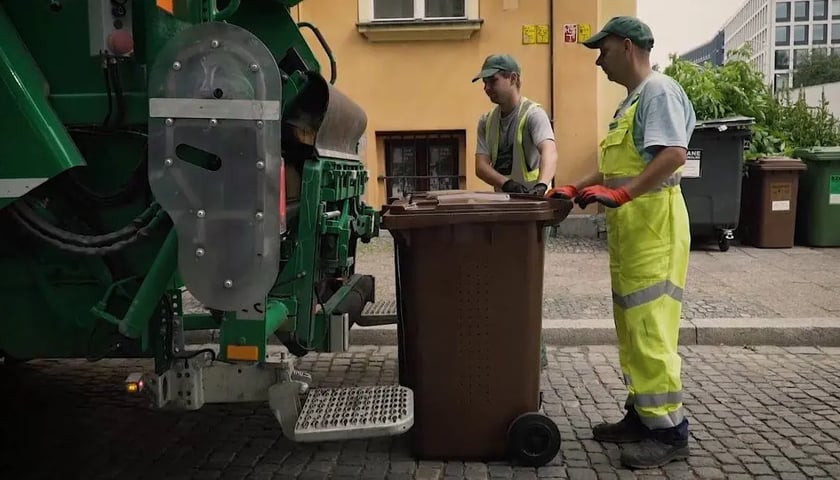 Na zdjęciu dwóch pracowników firmy wywożącej odpady prowadzi brązowy pojemnik na odpady do zaparkowanej śmieciarki. Zdjęcie ilustracyjne