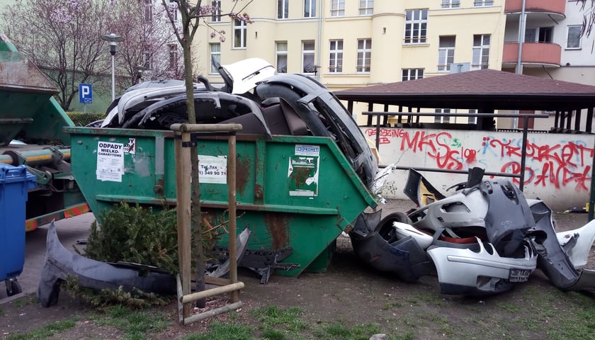 Zielony kontener na odpady wielkogabarytowe wypełniony częściami samochodu, które leżą także obok