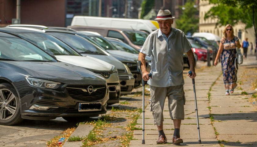 Powiększ obraz: Starszy mężczyzna o kulach idący chodnikiem w upalny dzień. Zdjęcie ilustracyjne
