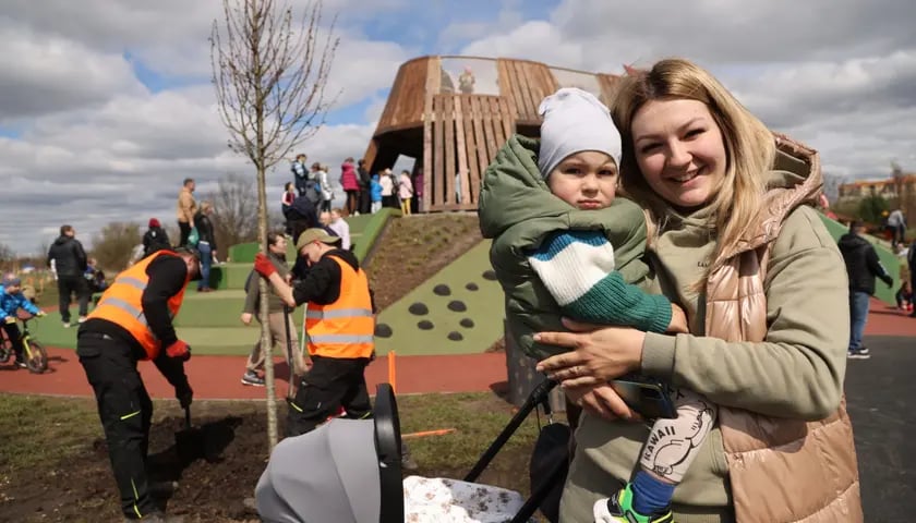 Uśmiechnięta kobieta trzyma dziecko, w tle trwa akcja WROśnij we WROcław i sadzenie drzewek w parku Tysiąclecia