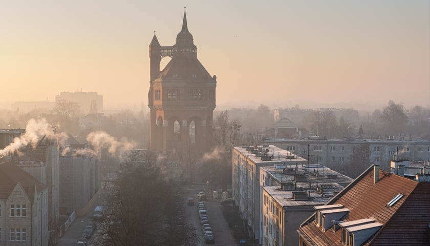 Na zdjęciu widać ulicę Sudecką, dachy budynków oraz wieżę ciśnień spowitą smogiem