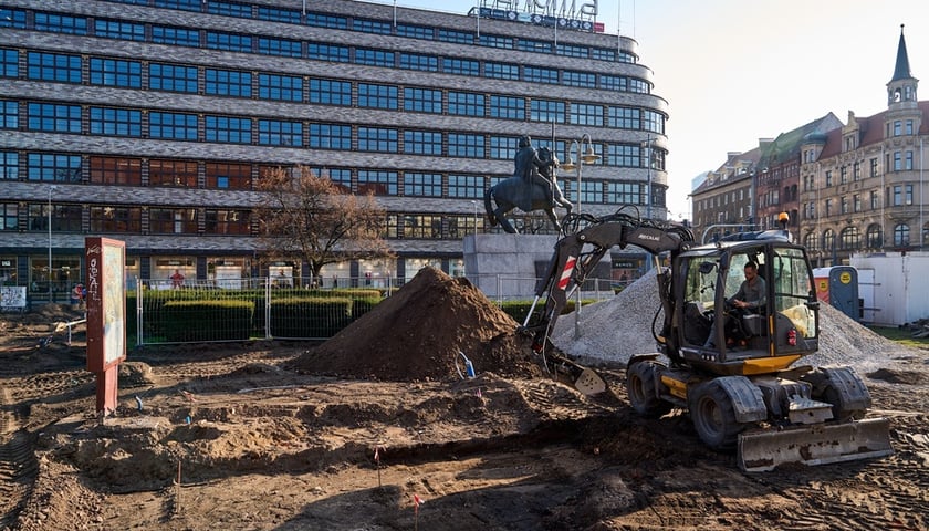Koparka z operatorem na wykopkach pod rośliny przy konnym pomniku Bolesława Chrobrego, na tle Renomy (kwiecień 2025)