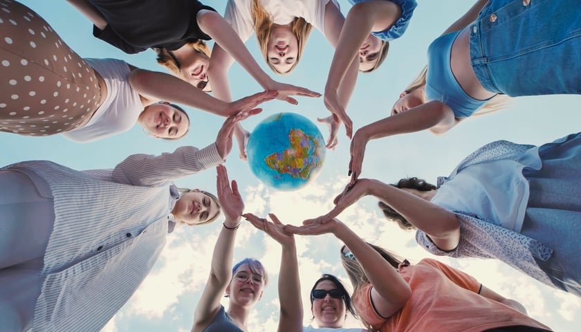 Grupa dzieci i młodzieży ustawionych w kole, fotografowana od dołu patrzących w górę na niebieskie niebo, z rękami wyciągniętymi w stronę kolorowego globu ziemskiego umieszczonego w centrum.