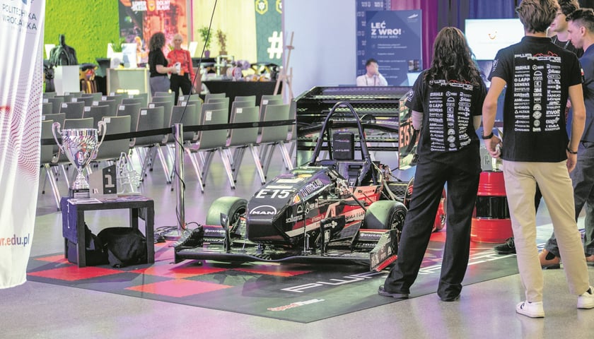 Model bolidu, po lewej stronie stoją puchary, po prawej dwoje młodych ludzi